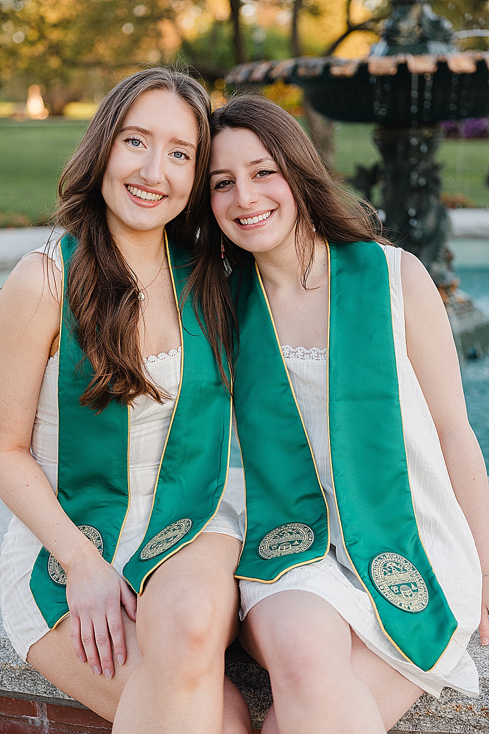 Two UVM senior friends smiling with the water fountain on the campus green behind them