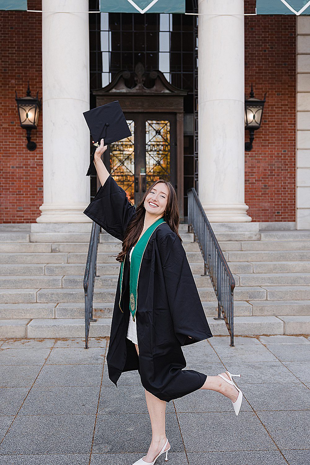 UVM senior girl smiling as she lifts her leg and her cap in front of the Waterman Building on campus